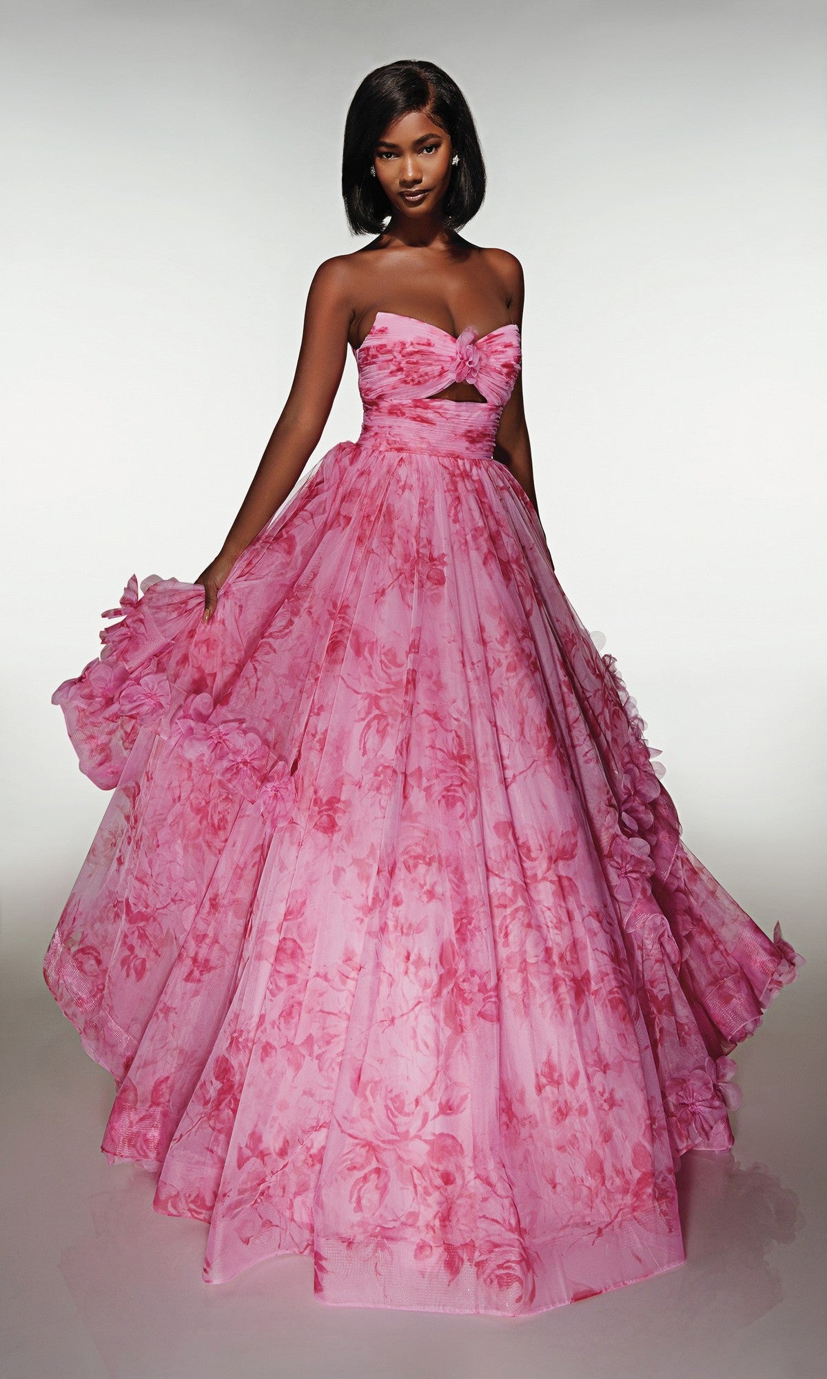 A woman poses against a plain light background in the Alyce 62042 Long Prom Dress in bubblegum, holding out the ruffled ballgown skirt to the side to showcase its strapless pink floral design.